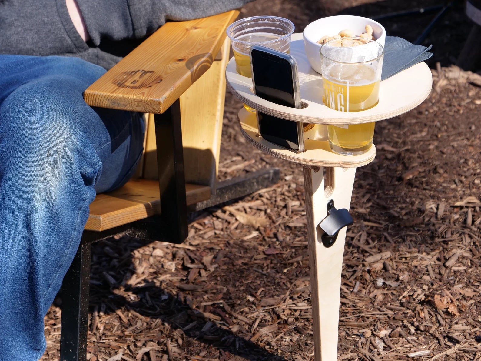 the light colored beer table holding two cups of beer, a phone, and bowl of snacks next to someone in a lawn chair