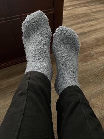 Person wearing fuzzy socks and black pants, with feet propped up on a wood floor, conveying a relaxed, cozy setting perfect for lounging or staying warm