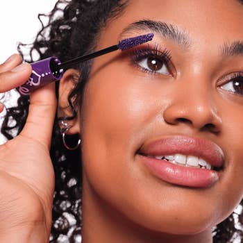 Person applying mascara with a purple wand, smiling and looking upwards, highlighting lashes