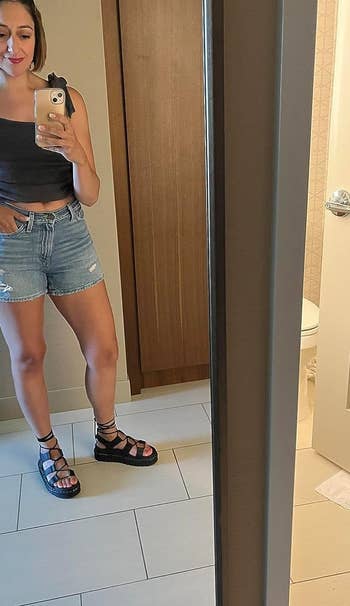 reviewer taking a mirror selfie, wearing a casual one-shoulder top and denim shorts, paired with strappy sandals