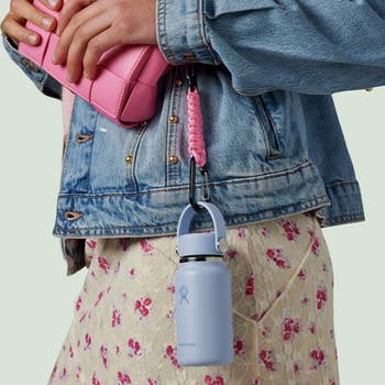 Person with denim overgarment   holds a pinkish  bag, with a lavender h2o  vessel  clipped on. They're wearing a floral skirt