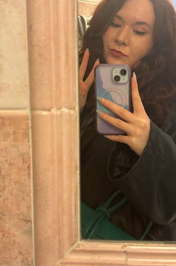 Person taking a mirror selfie holding a phone, wearing lipstick and a dark jacket