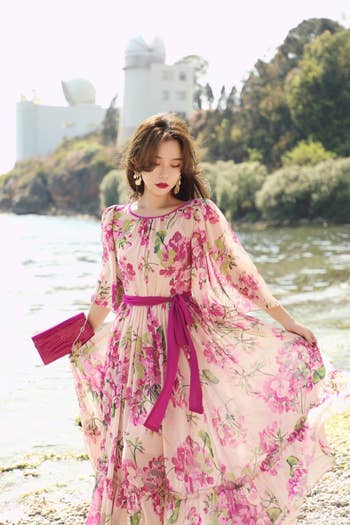 model in a floral dress with a purple belt and clutch, standing by the water
