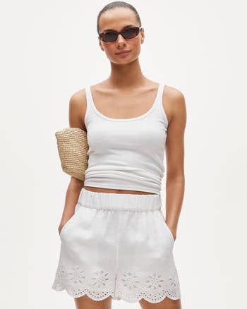 Model wearing a sleeveless apical and eyelet shorts, accessorized with sunglasses and a woven clutch