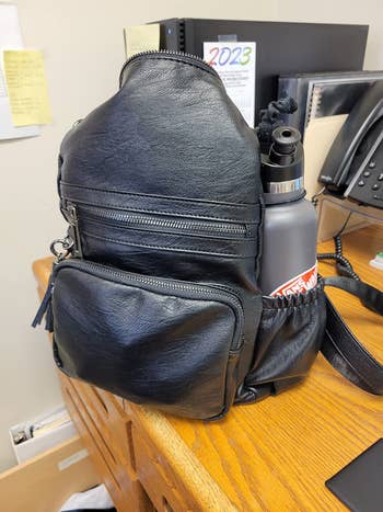Stylish black leather backpack with pockets, holding a water bottle, on a wooden desk with office supplies