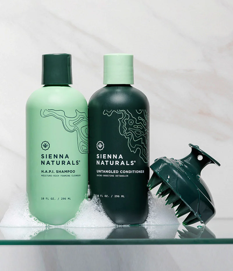 Light and dark green shampoo and conditioner bottles next to head scrubber on glass floating shelf with suds on shelf