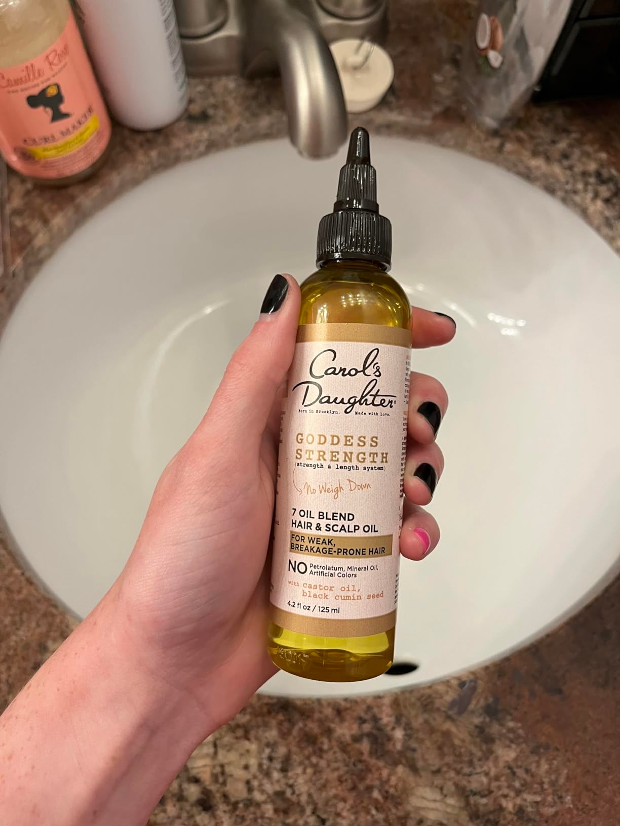 Hand holding a bottle of Carol's Daughter Goddess Strength Hair & Scalp Oil near a sink