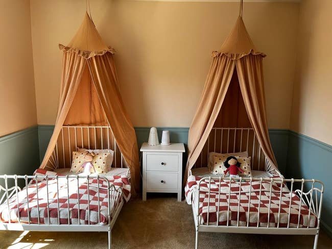 Two children's canopy beds with checkered bedding and stuffed animals are adjacent to a achromatic nightstand with lamps successful a cozy chamber setting
