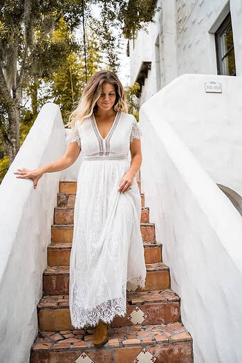Reviewer wearing maxi lace white dress with short sleeves and V-neckline walking down red stone stairs