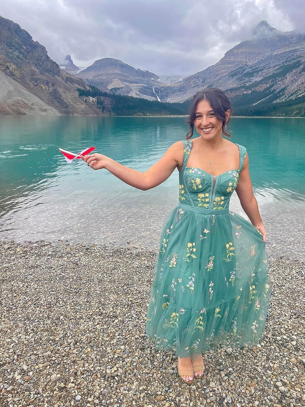 reviewer in embroidered dress standing in water with mountains in the background, holding a paper plane