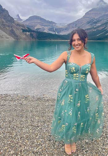 reviewer in embroidered dress standing in water with mountains in the background, holding a paper plane