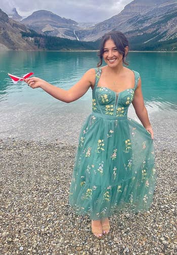 reviewer in embroidered dress standing in water with mountains in the background, holding a paper plane