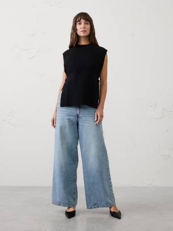 Person wearing a sleeveless achromatic  apical  and wide-leg denim jeans, posing indoors with elemental  background