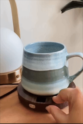 A manus  adjusts a mug warmer with a ceramic mug connected  it, suggesting a cozy, convenient setup for keeping drinks hot