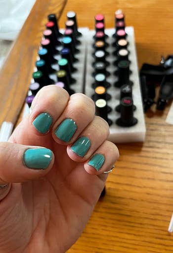 Person showing hand with teal nail polish, selection of various nail polish bottles in the background
