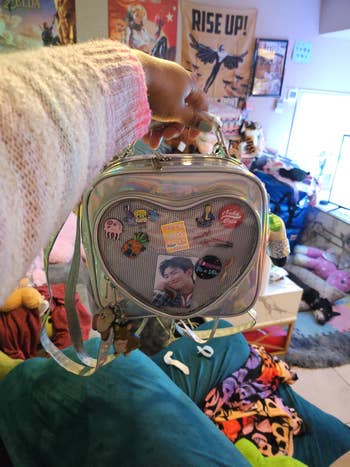 Person holding a clear heart-shaped backpack adorned with various pins, including a celebrity photo. Background shows a colorful, cozy room