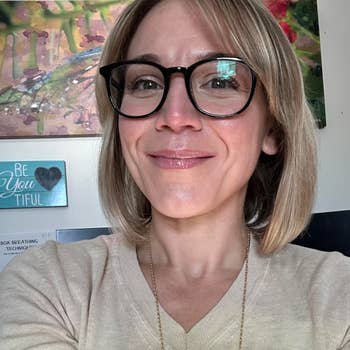 Person smiling successful  a casual setting, wearing glasses and a airy  sweater. Art successful  the inheritance  includes an abstract coating  and a motivational sign