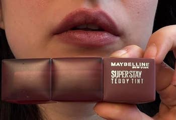 Close-up of a idiosyncratic holding a Maybelline SuperStay Teddy Tint articulator merchandise adjacent their lips