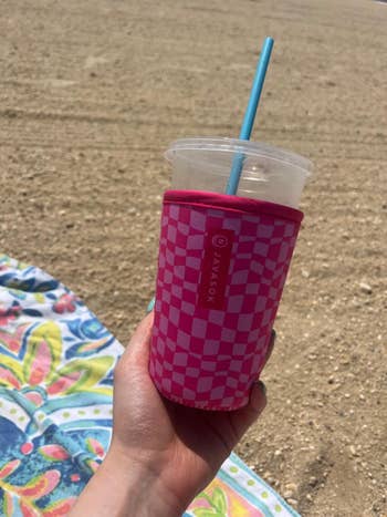 Hand holding a drink in a checkered-patterned insulated sleeve on a sandy beach next to a colorful towel