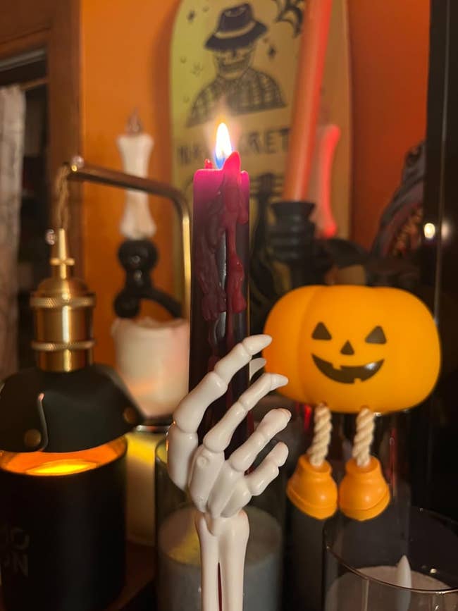 A skeleton manus  candle holder grips a lit, acheronian  candle beside Halloween decor, including a jack-o'-lantern light