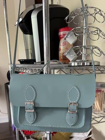 Light blue satchel with buckle closures hanging on kitchen shelf, featuring a coffee maker, creamer, and coffee pods in background