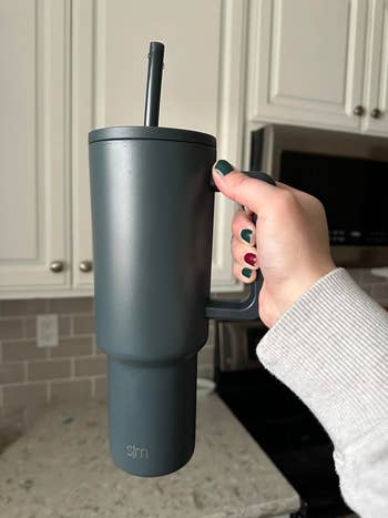 Person holding a large, insulated travel mug with straw in a kitchen setting