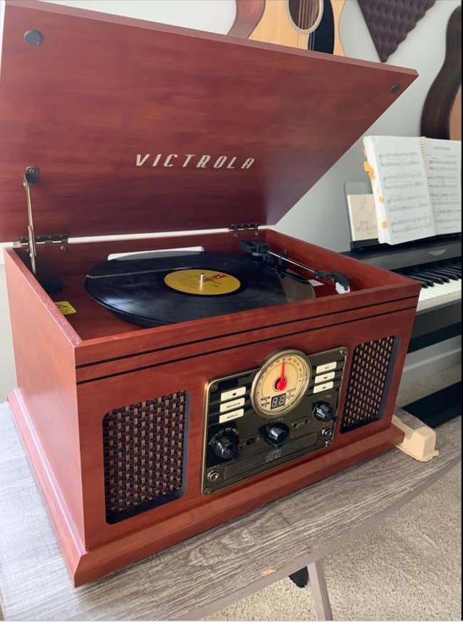 Victrola record player with a vinyl record playing. Background includes an open sheet music book on a piano, and several guitars hanging on the wall