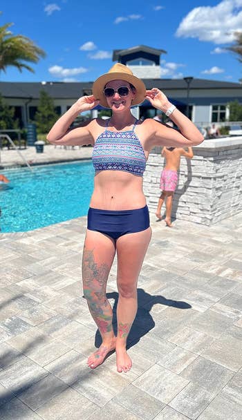 Person in a swim top and hat by a poolside, showing floral tattooed leg, smiles and poses with hands on hat brim