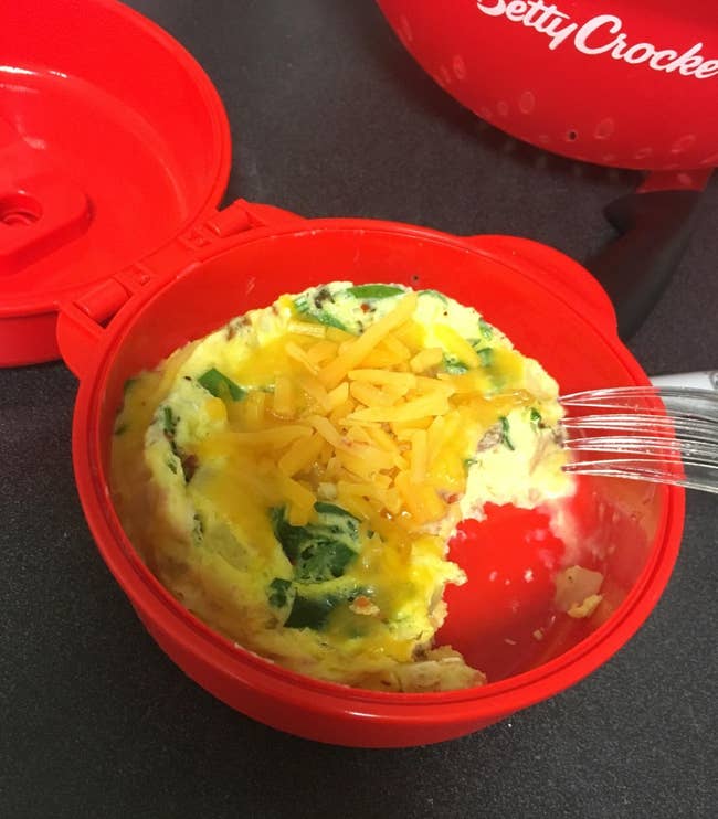 A small red circular cooker with an egg with veggies and cheese in it 