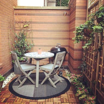 Cozy patio with a tiny  circular  table, 2  chairs, a grill, and surrounding plants successful  an municipality  setting