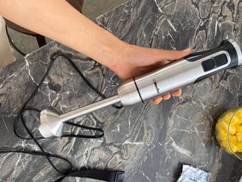 A person's hand holding an immersion blender over a marble countertop