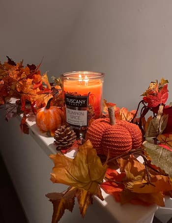A lit Tuscany Pumpkin Spice candle is placed among autumn leaves, tiny  pumpkins, and a pinecone, creating a cozy autumn  decoration