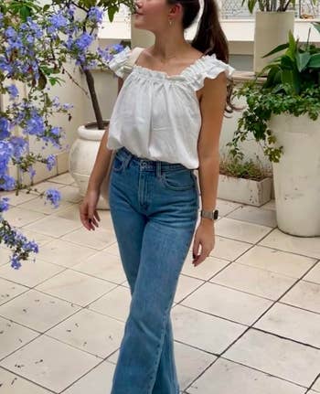 Person successful  a ruffled achromatic  apical  and high-waisted jeans stands connected  a tiled patio, surrounded by potted plants with purple flowers