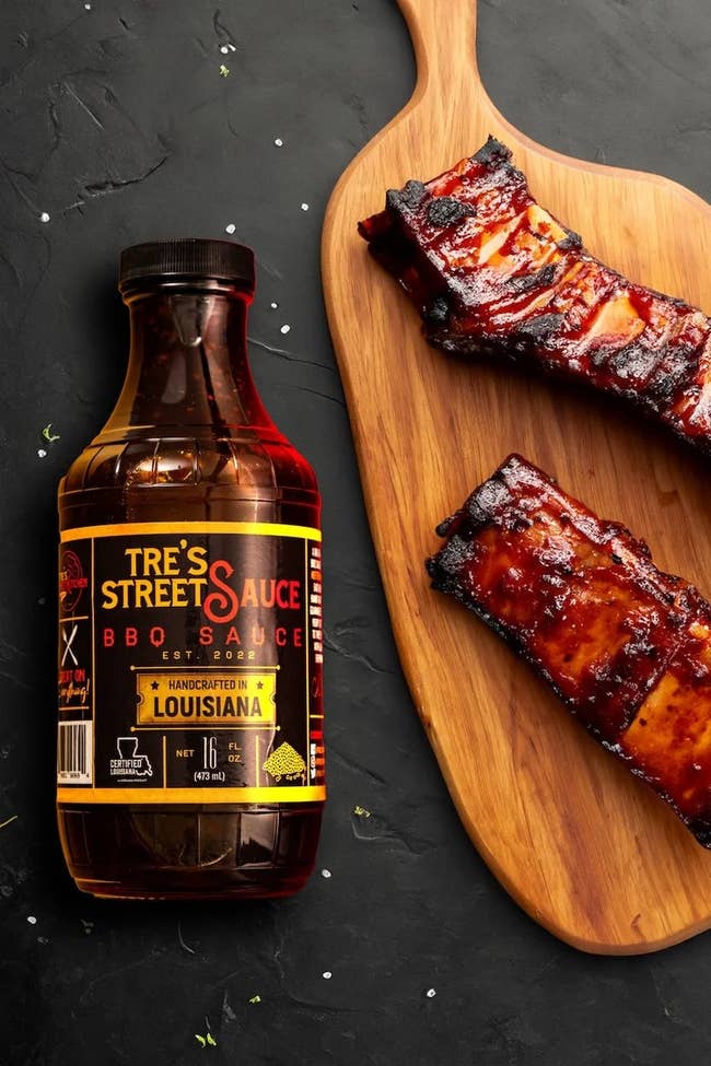 Bottle of Tre's Street Sauce BBQ Sauce adjacent to glazed ribs connected a woody board, acceptable against a acheronian background
