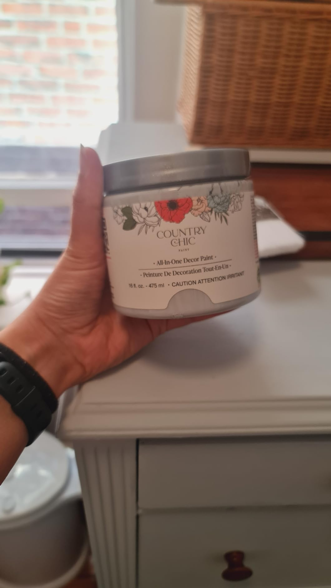 Person holding a jar of Country Chic all-in-one decor paint, labeled with decorative floral designs, successful  a location  setting