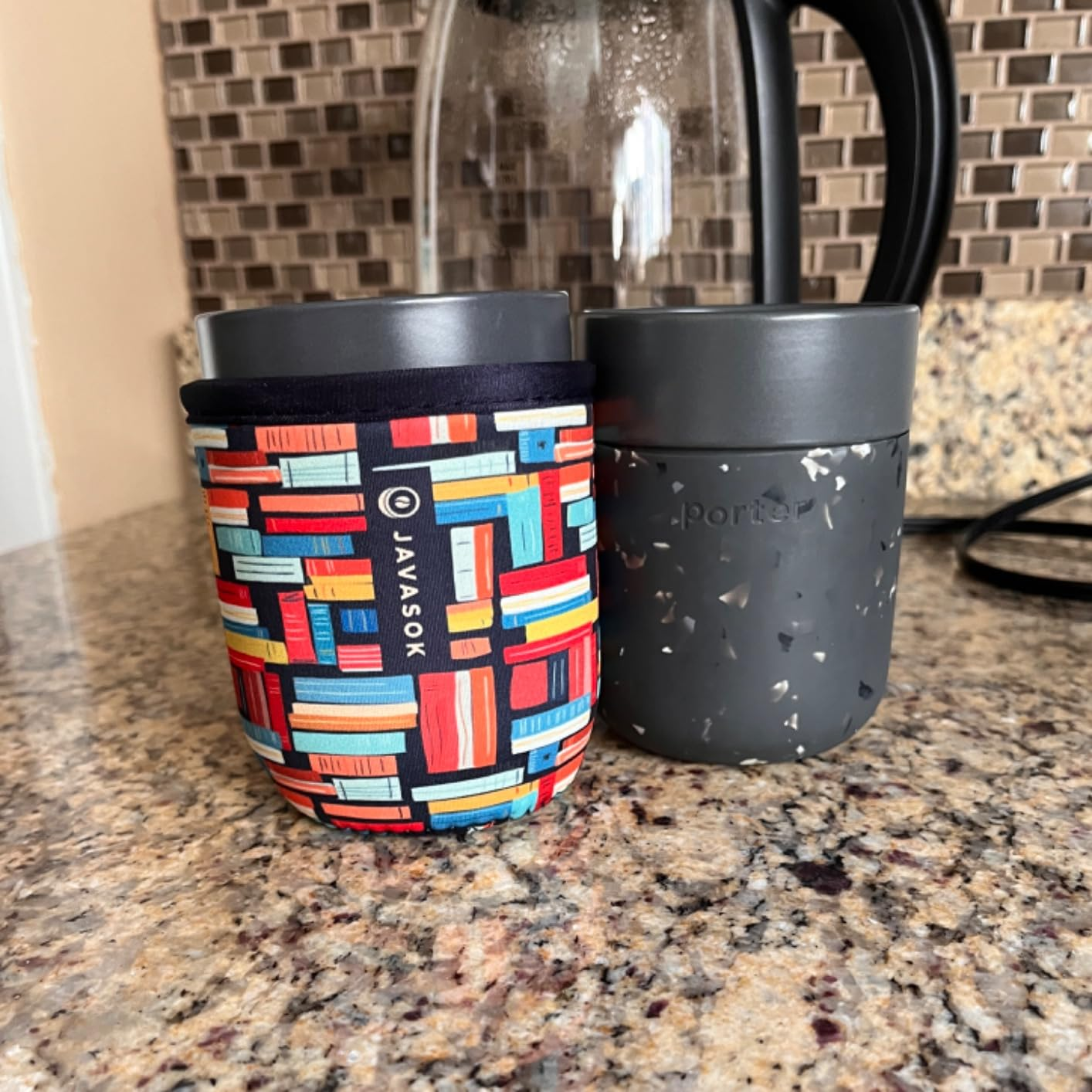 Two reusable cups on a counter; one has a colorful geometric sleeve, and the other is solid, with a speckled design