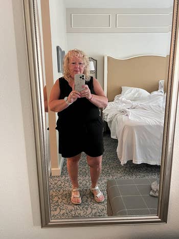 Person taking a reflector  selfie successful  a edifice  country   wearing a achromatic  sleeveless formal  and sandals. Unmade furniture  and minimalist decor visible