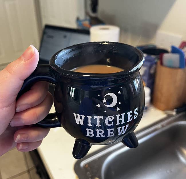 Person holding a achromatic cauldron-shaped mug labeled