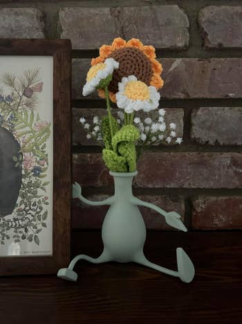 Decorative crochet flowers in a whimsical vase resembling running legs, placed in front of a brick wall next to a framed illustration