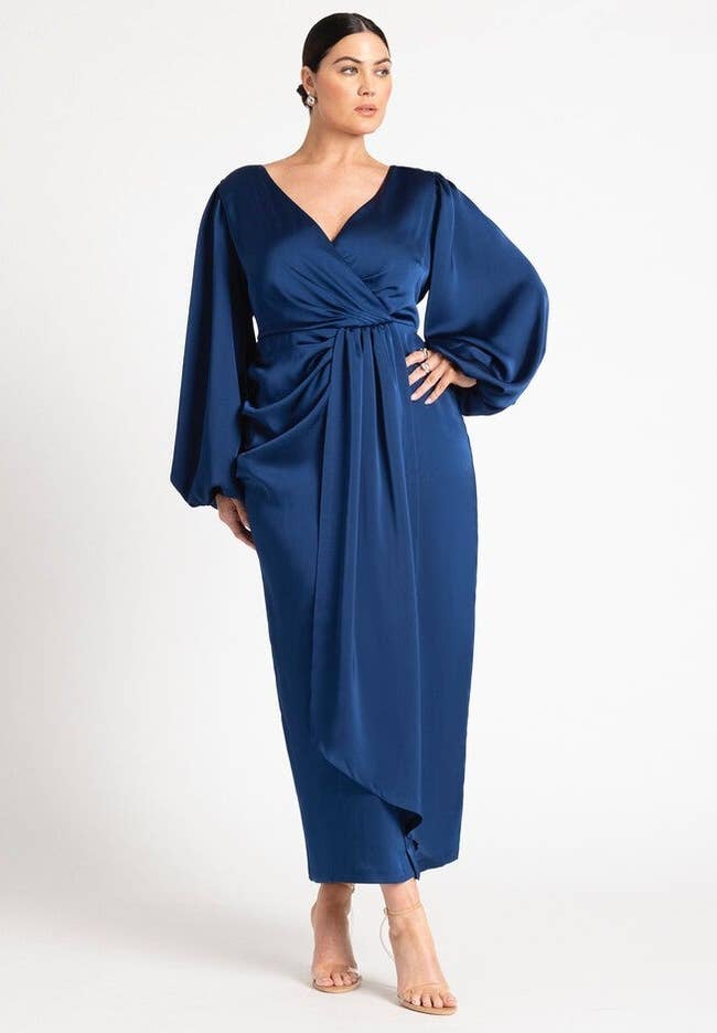 Person wearing an elegant, long-sleeved, blue gown with a gathered front detail, standing in a fashion pose