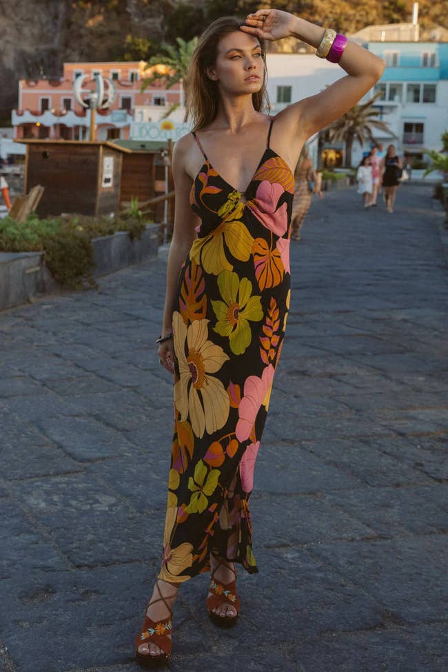 Model wearing black sleeveless maxi dress with yellow, green, and pink flower pattern and deep sweetheart neckline paired with brown strappy sandals on stone ground