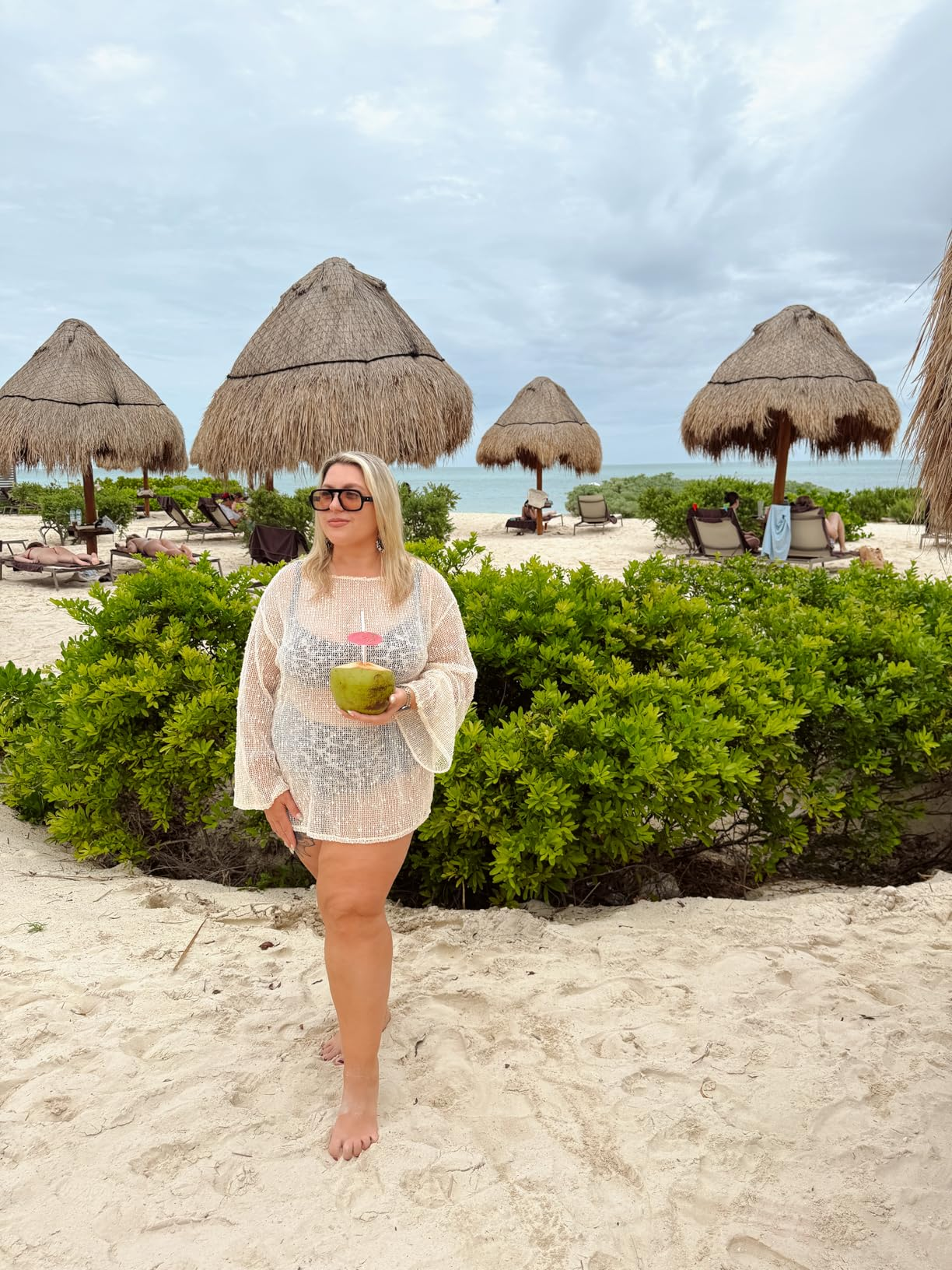 Person connected  sandy formation  holding a coconut drink, wearing a sheer cover-up, with thatched umbrellas and lounge chairs successful  the background