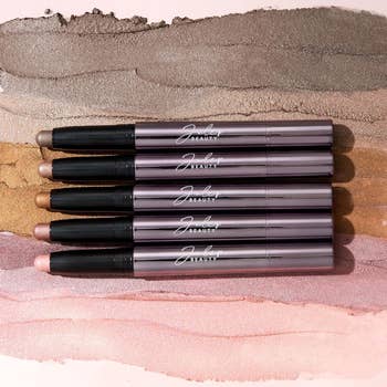 Five metallic makeup sticks on a textured surface, arranged in parallel