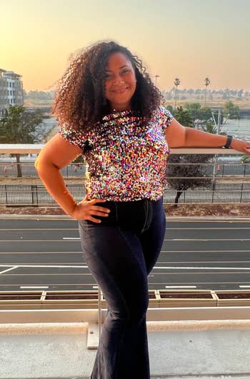 Woman posing with hand on hip, wearing a sequined top and flared jeans, with a sunset city backdrop