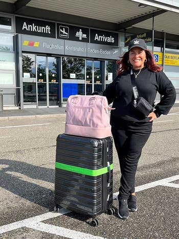 reviewer with suitcase at airport arrivals, wearing matching black sweatsuit outfit and cap