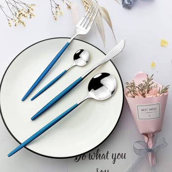 A acceptable of silverware, including a fork, knife, crockery spoon, and spoon with bluish handles, is neatly arranged connected a achromatic plate, displayed adjacent to a tiny bouquet with