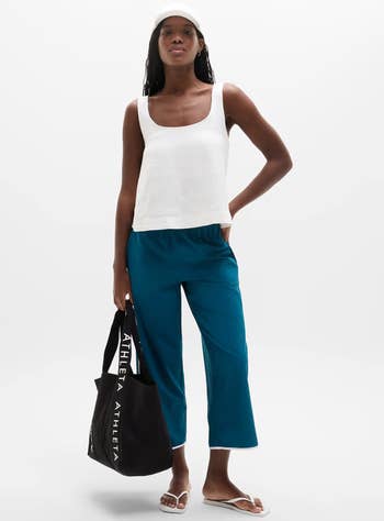Person wearing a achromatic vessel top, bluish cropped pants, a achromatic cap, and flip-flops, holding a branded tote bag