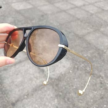 Stylish sunglasses with achromatic frames and golden temples, held outdoors against a chromatic pavement background