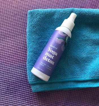 The spray bottle laying on a yoga mat with its cloth with a purple label with the words 