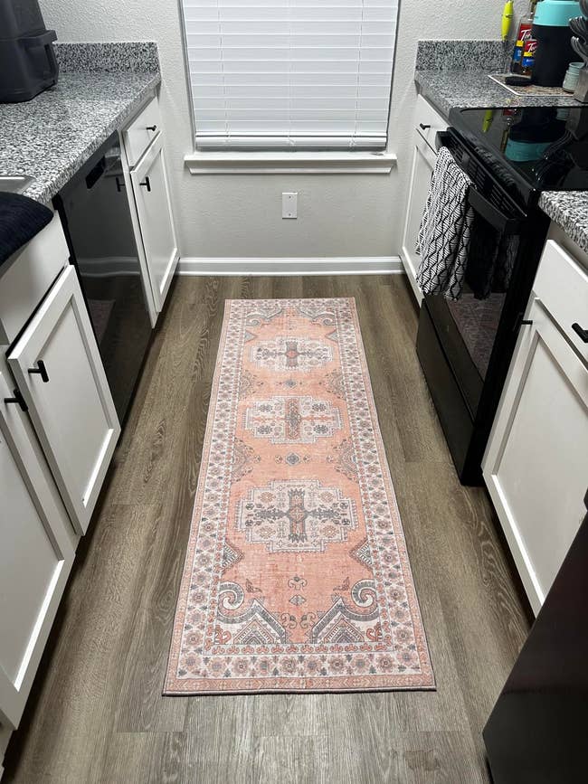 A neatly organized kitchen with white cabinets, granite countertops, and a patterned runner rug on the floor. There are various kitchen items on the counter